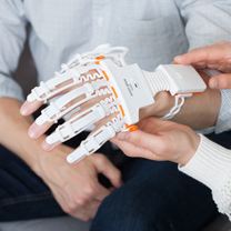 Pacjent ma ubraną rękawicę Neofect na lewej dłoni. Tą rękę podtrzymuje dłoń kobiety.